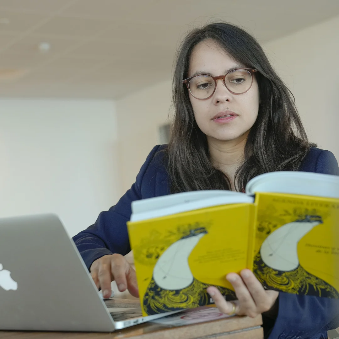 Mujer con un libro en la mano tomando notas en el ordenador